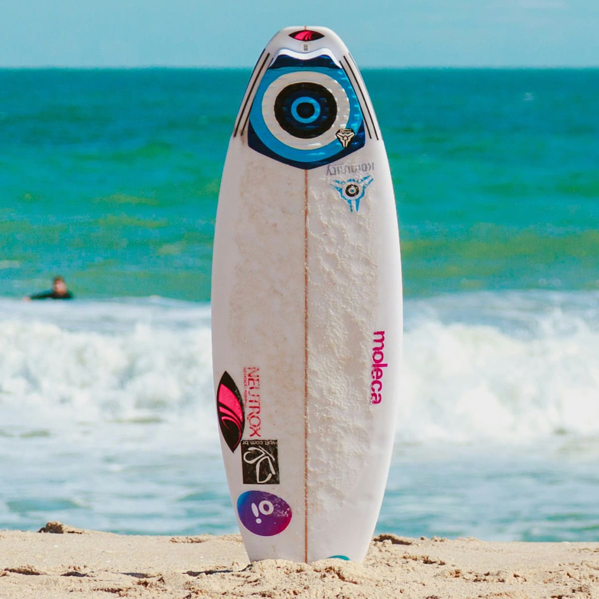 surfboard stuck in sand upright on beach with waves and a surfer in the water in the background